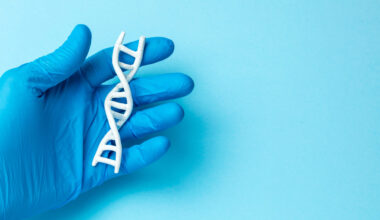 imagem de fundo azul claro, com uma mão enluvada por luva cirúrgica também azul, mas mais escuro, segurando um molde de DNA