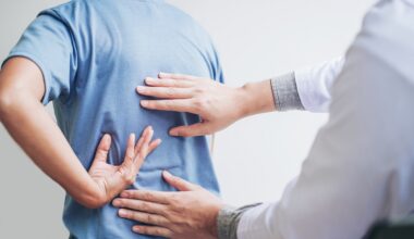 médico realizando atendimento em paciente com aparente dor nas costas. paciente está de blusa azul, apontando para o local de dor e médicos com as mãos sobre o mesmo